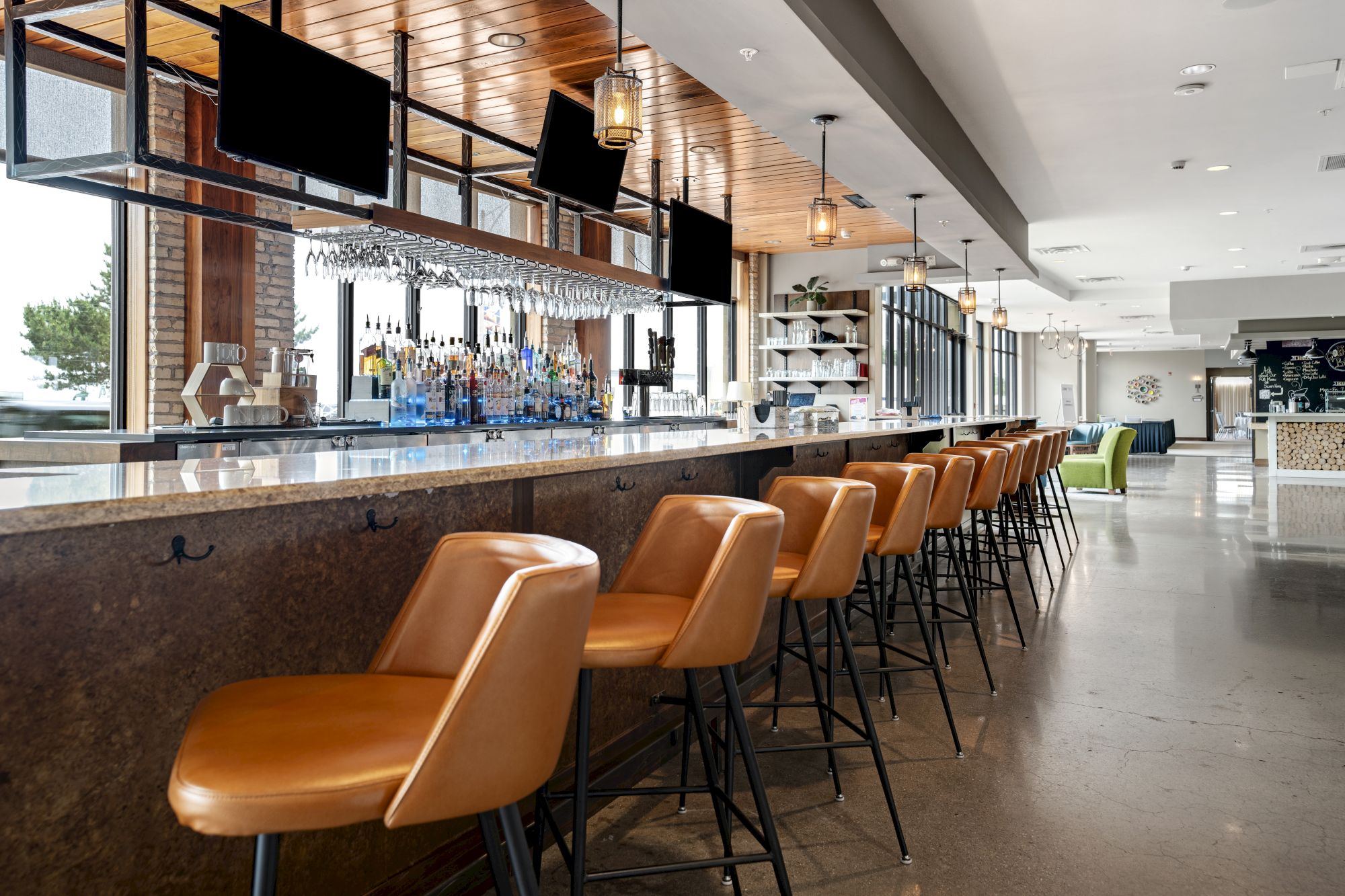 A modern bar with a long counter, tan stools, overhead screens, hanging lights, and a display of various bottles and glasses.