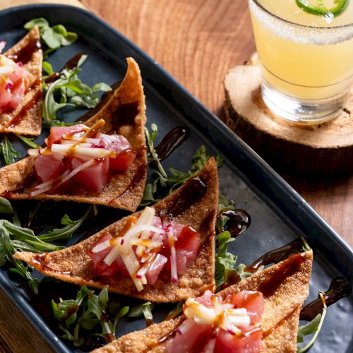 Crispy wonton chips topped with diced tuna and garnish on greens beside a cocktail with lime and jalapeño slices.