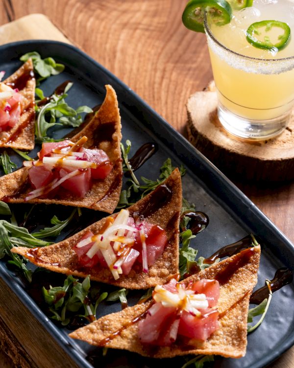 Crispy wonton chips topped with diced tuna and garnish on greens beside a cocktail with lime and jalapeño slices.