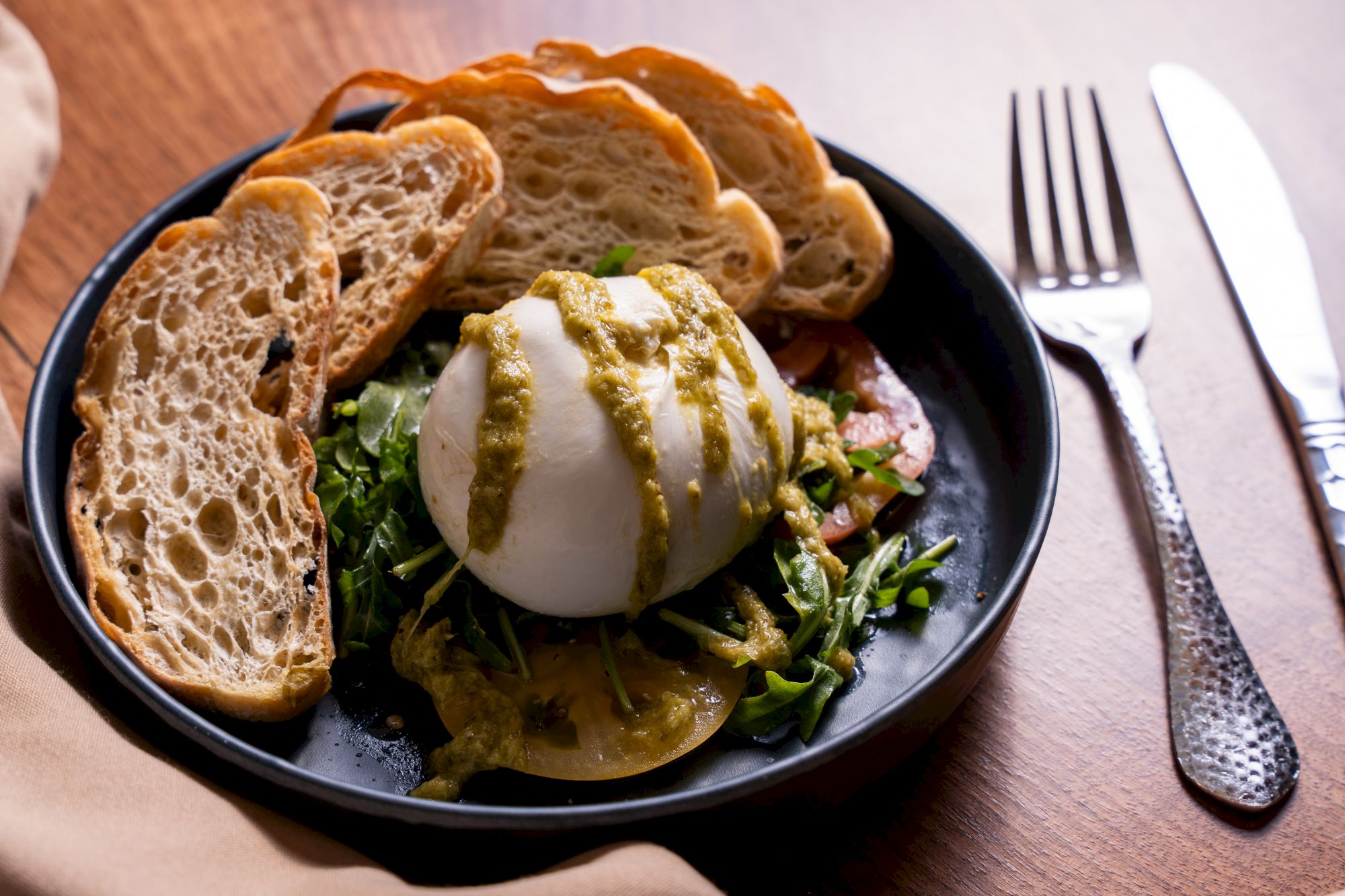 A plate with burrata covered in green sauce, surrounded by arugula, tomatoes, and sliced bread, fork and knife beside it.