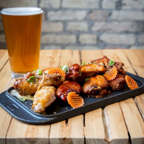 A tray of chicken wings garnished with herbs and crisps is next to a glass of beer on a wooden table with a brick wall backdrop.