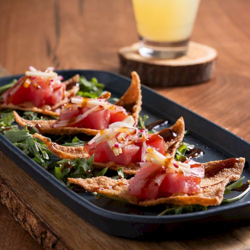 The image shows a dish with sliced tuna on crispy wontons, garnished with greens, and a cocktail in the background on a wooden table.