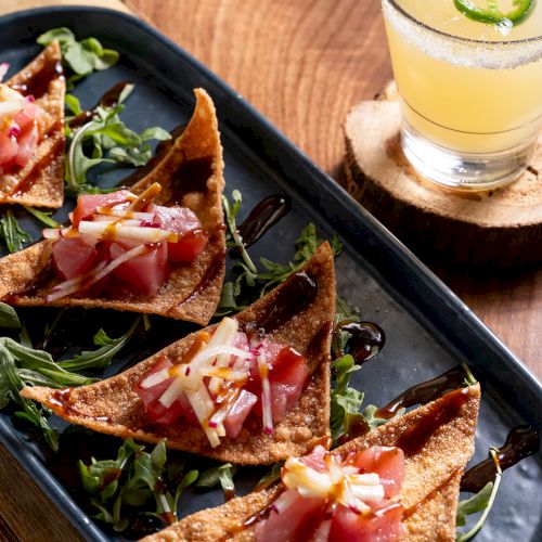 The image shows a plate with crispy tostadas topped with diced tuna and cheese, accompanied by a cocktail garnished with jalapeño slices.