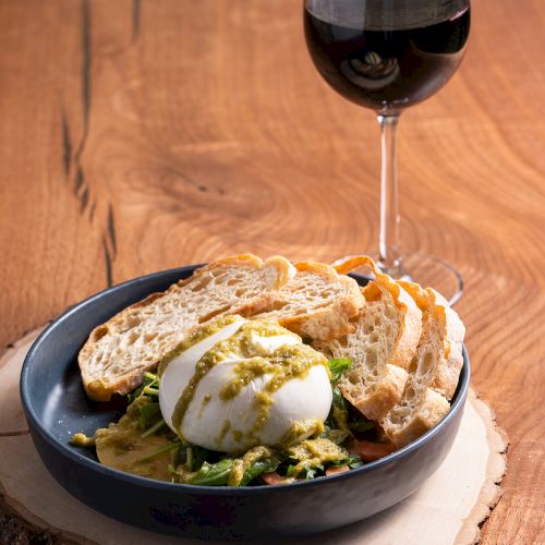 A bowl with bread slices, a poached egg on greens, and a glass of red wine on a wood slice serving as a placemat.