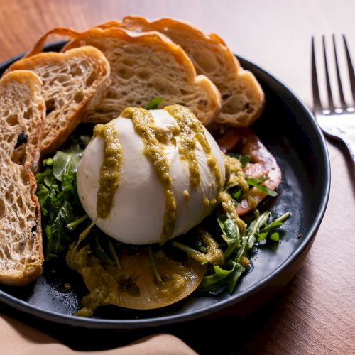 A plate with burrata cheese, pesto, arugula, and tomato slices, accompanied by toasted bread slices, setup beside a fork and knife.