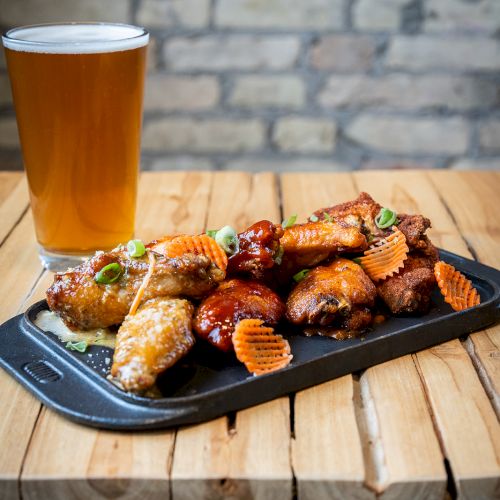 A glass of beer sits on a wooden table next to a tray of assorted chicken wings, garnished with herbs and waffle-cut fries.