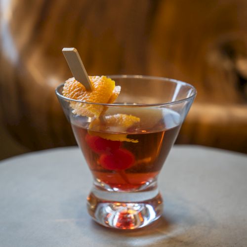 A cocktail glass with a dark drink, garnished with citrus peel and a cherry, sits on a table in a cozy setting.