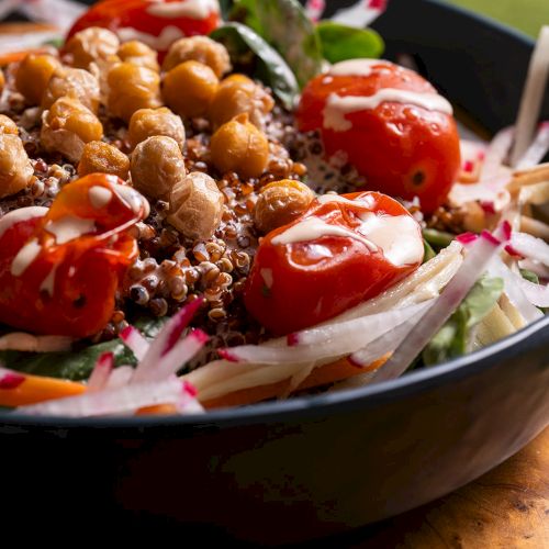 A fresh salad with chickpeas, cherry tomatoes, quinoa, and mixed greens in a black bowl, garnished with sliced radishes and onions.