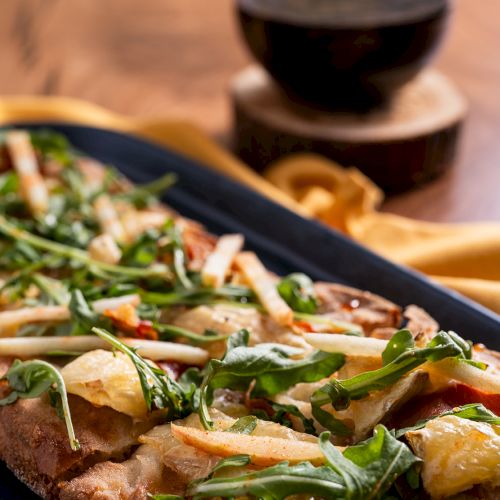 A flatbread topped with arugula and other ingredients served on a platter, with a glass of red wine in the background on a wooden table.