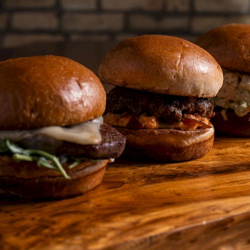 The image shows three burgers with different toppings on a wooden surface, set against a brick wall background.