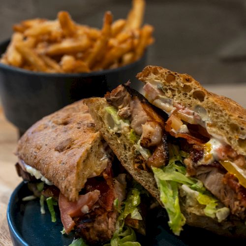 A sandwich with lettuce and tomato on a plate, and a bowl of fries in the background against a dark-toned setting.