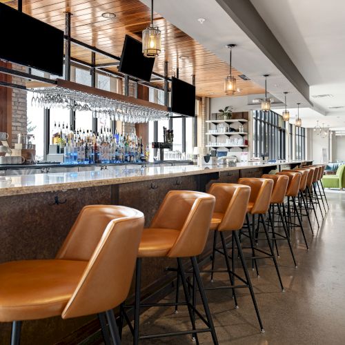 A modern bar with tan leather stools, a marble countertop, hanging glassware, bottles, and black screens, with seating visible in the distance.