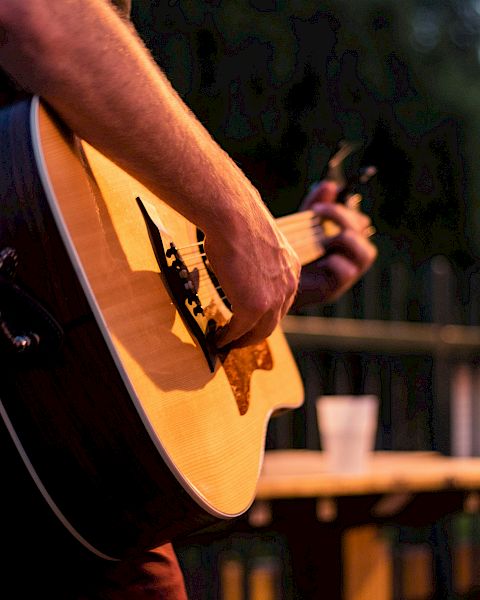 A person playing an acoustic guitar outdoors with a blurred background, likely in the evening, near a table with a cup.