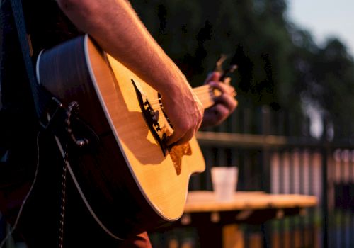 A person playing an acoustic guitar outdoors with a blurred background, likely in the evening, near a table with a cup.