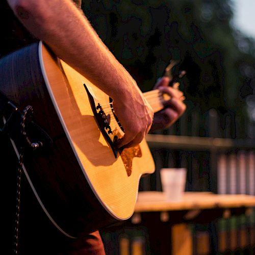 A person playing an acoustic guitar outdoors with a blurred background, likely in the evening, near a table with a cup.