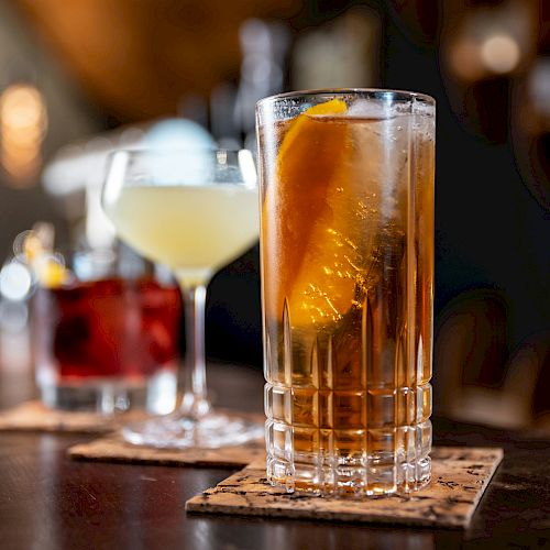 The image shows three cocktails on a bar: a tall glass with an orange slice, a coupe glass with a light drink, and a short glass with ice.