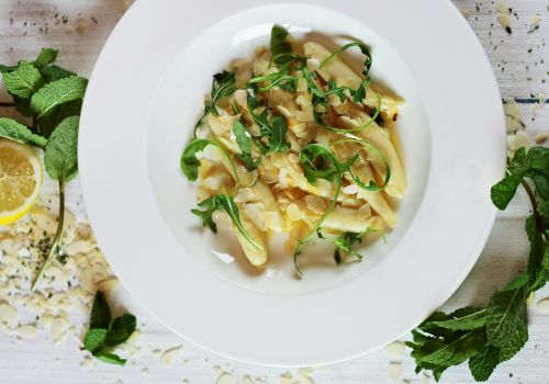 A plate of pasta topped with fresh greens and herbs, surrounded by mint leaves, sliced almonds, and a lemon wedge on the side.