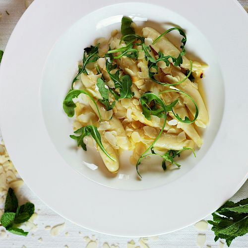 A plate of pasta topped with fresh greens and herbs, surrounded by mint leaves, sliced almonds, and a lemon wedge on the side.